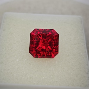 May include: A faceted, radiant-cut, red gemstone. The gem is a deep, vibrant red color and is set against a white background. The gemstone has a square shape with beveled edges, reflecting light.