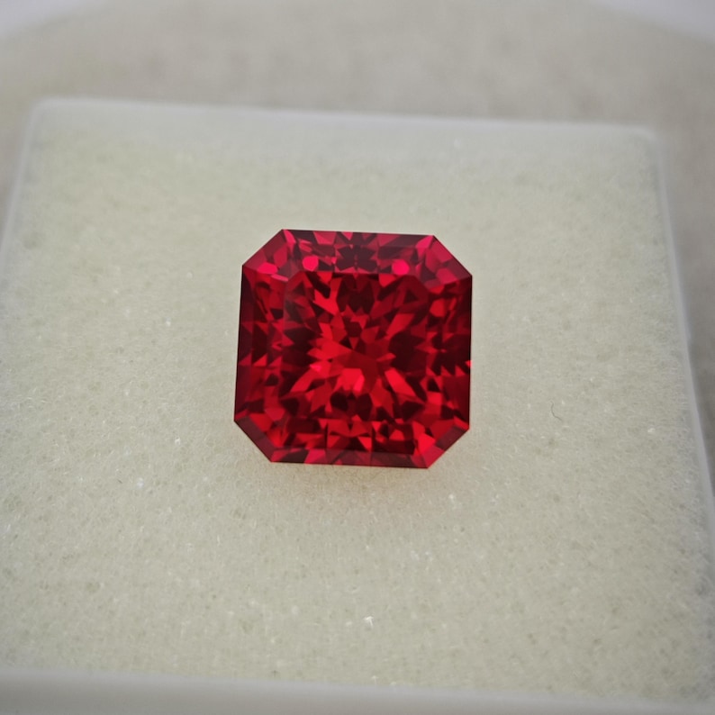 May include: A faceted, radiant-cut, red gemstone. The gem is a deep, vibrant red color and is set against a white background. The gemstone has a square shape with beveled edges, reflecting light.