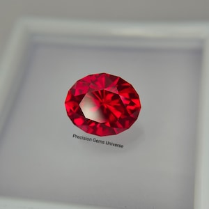 May include: A vibrant, round-cut red gemstone with multiple facets that reflect light, creating a brilliant display of colour. The text Precision Gems Universe is printed in black below the stone, which is set against a plain white background.