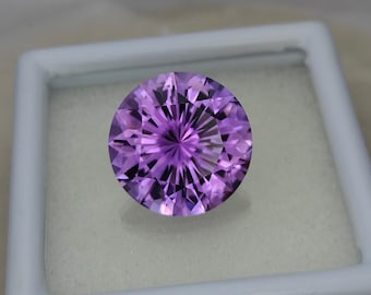 Könnte beinhalten: Ein runder, facettierter Amethyst-Edelstein mit einer violetten Farbe. Der Edelstein ist in einer weißen Schachtel platziert.