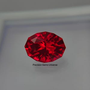 Puede incluir: Una gema roja de corte ovalado con facetas intrincadas que crean una superficie brillante y reflectante. La piedra muestra un tono carmesí profundo y saturado en toda su estructura. El texto Precision Gems Universe aparece impreso debajo.
