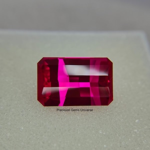 May include: A rectangular, faceted gemstone with a deep red hue. The gem has a bright pink interior and is labeled "Precision Gems Universe". The cut of the stone creates multiple planes that reflect light.