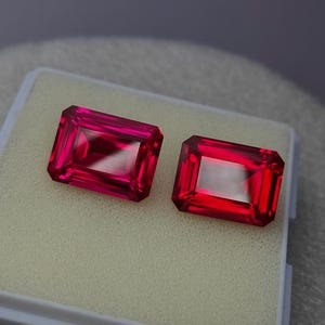 Könnte beinhalten: Zwei rechteckige, facettierte, rote Edelsteine. Die Edelsteine werden nebeneinander in einem weißen, schaumstoffgefütterten Etui präsentiert. Die Edelsteine haben ein lebendiges, glänzendes Aussehen mit einem klaren, reflektierenden Inneren.