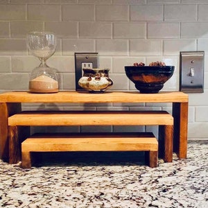 Puede incluir: Un conjunto de tres elevadores de madera de diferentes tamaños, ideal para exhibir objetos. Los tonos de madera natural complementan los objetos decorativos, incluyendo un reloj de arena de vidrio, una olla de cerámica y un cuenco de madera oscura.