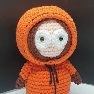Puede incluir: Un juguete de peluche tejido a crochet de un personaje de dibujos animados que lleva una chaqueta con capucha naranja con ribetes negros. El personaje tiene el pelo marrón, los ojos blancos y la cara rosa.