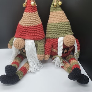 Op de afbeelding: Twee gehaakte gnomen die naast elkaar zitten. De gnome links draagt een rode en bruine muts met een witte baard en zwarte laarzen. De gnome rechts draagt een groene en bruine muts met een witte baard en zwarte laarzen. Beide gnomen hebben gouden belletjes op hun mutsen.