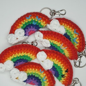May include: Five crocheted rainbow keyrings with white clouds and silver key rings. Each keyring features a rainbow with red, orange, yellow, green, blue, indigo, and violet colours.