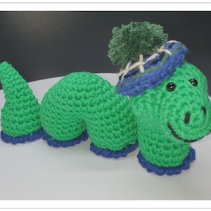 May include: A green crocheted caterpillar wearing a blue and white plaid hat with a green pom-pom. The caterpillar has a smiling face with black eyes and a black mouth.