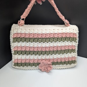 May include: A handmade crochet handbag in cream, pink, and green stripes, with a matching pink flower accent and handle. The bag is rectangular with a flap closure and metal rings attaching the handle.