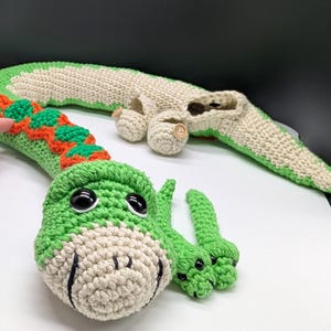 May include: Crocheted green and cream-colored dragon toy with orange accents. The dragon has large black eyes and a smiling face. The dragon's body is segmented, and the tail is long and curved.