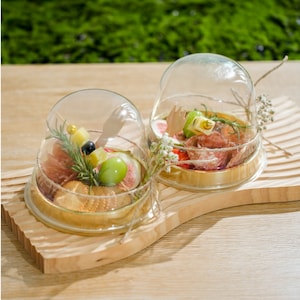 May include: Two glass cloche domes with wooden bases, filled with small snacks. The left dome contains crackers, cheese, and fruit. The right dome contains salami, cheese, and fruit.