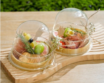 May include: Two glass cloche domes with wooden bases, filled with small snacks. The left dome contains crackers, cheese, and fruit. The right dome contains salami, cheese, and fruit.