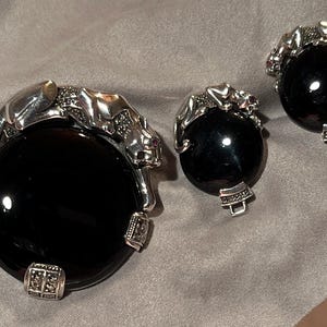 May include: A jewelry set featuring a large, round black stone pendant and matching earrings. Each piece is adorned with a silver panther design, with red accents on the eyes. The set is displayed on a gray fabric surface.