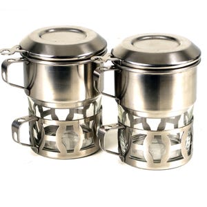 Peut inclure: Deux filtres à café vietnamiens en acier inoxydable, chacun composé d'une chambre cylindrique avec couvercle et poignée, reposant dans une tasse en verre transparent maintenue par un cadre métallique décoratif ajouré avec une poignée latérale.
