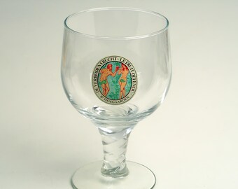 May include: A clear glass beer goblet with a round bowl and a twisted stem. The glass has a circular emblem with two figures and text in a circle. The text includes the words "DE VERBODEN VRUCHT" and "LE FRUIT DE FENDU".