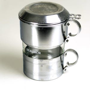 Op de afbeelding: Een Vietnamese koffiefilter van roestvrij staal, met een glazen beker en het woord "DUROBOR" op het glas gedrukt. De filter heeft een deksel en twee handvatten. De koffiefilter is ontworpen om koffie te zetten.