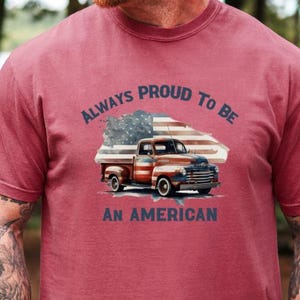 May include: Maroon t-shirt featuring a graphic of a vintage pickup truck with an American flag design. The text reads "ALWAYS PROUD TO BE AN AMERICAN" in navy blue. The shirt is a short-sleeved crew neck.