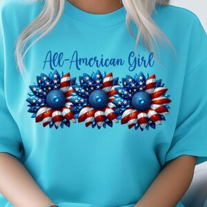 May include: A light blue t-shirt featuring three sunflower designs with red, white, and blue patterns, resembling the American flag. The text "All-American Girl" is arched above the flowers in blue script.
