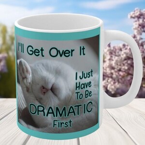 May include: White ceramic mug with a teal blue background and a white cat sleeping on it. The text on the mug reads "I'll Get Over It I Just Have To Be Dramatic First."