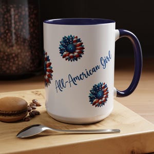 May include: White ceramic mug with a blue interior and handle. The mug features three sunflower designs in red, white, and blue, with the text "All-American Girl" in blue script. The mug is on a wooden surface.