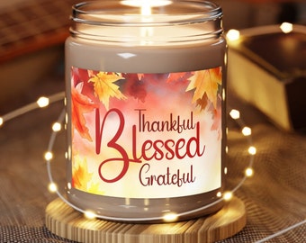 May include: A brown glass candle with a white label featuring a watercolor design of autumn leaves and the text "Thankful Blessed Grateful". The candle is lit and surrounded by white string lights.