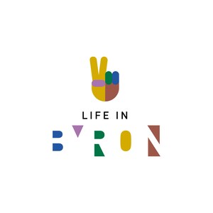 Op de afbeelding: Een kleurrijk grafisch ontwerp met de tekst "LIFE IN BYRON" in zwarte letters. De "B" is blauw, de "Y" is paars, de "R" is groen, de "O" is geel en de "N" is bruin. De grafiek toont ook een gele hand die een vredesteken maakt met een paarse band, groene en blauwe vingers en een bruine pols.