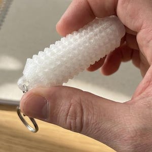 May include: A translucent white, cylindrical keyring featuring a repeating, textured, spiked geometric pattern across its entire surface. The object is attached to a small, circular silver-coloured metal split ring at one end for secure attachment.