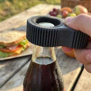 May include: A black bottle opener removing a bottle cap from a glass bottle. The opener has a ridged circular design and is held over the bottle. A sandwich and fruit are in the background.