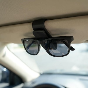 Könnte beinhalten: Schwarze Sonnenbrille an einem schwarzen Sonnenblendenclip befestigt. Die Sonnenbrille hat einen rechteckigen Rahmen und dunkle Gläser. Der Clip ist am Innenhimmel eines Autos befestigt und bietet eine praktische Aufbewahrungslösung für Brillen.