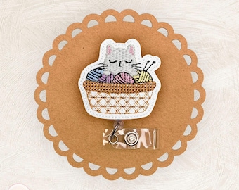 May include: A white and gray cat badge reel with a basket of yarn. The cat is embroidered with closed eyes, and the basket contains colorful yarn balls and knitting needles. The badge reel is attached to a brown scalloped circle.