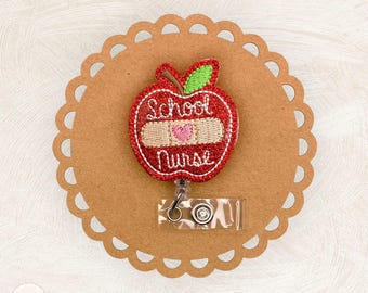 May include: A red apple-shaped badge reel with the words "School Nurse" embroidered in white. The apple has a bandage with a pink heart. The badge reel is on a brown scalloped circle.