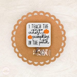 May include: A white square badge reel with rounded corners featuring black embroidered text that reads I TEACH THE CUTEST PUMPKINS IN THE PATCH, accented with two small orange embroidered pumpkins, attached to a clear plastic belt clip mechanism.