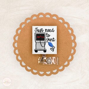May include: Embroidered badge with the text "Just need to vent" and a medical ventilator design. The badge is white with black, gray, red, and blue accents. It is attached to a clear plastic badge holder with a metal clip. The badge is on a brown, scalloped-edge background.
