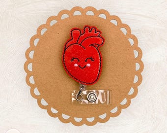 May include: A red, smiling heart-shaped badge reel with black stitching and a glittery finish. The badge reel is attached to a clear plastic holder with a metal clip. The badge is on a brown, scalloped-edge background.
