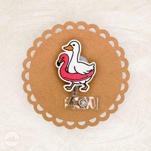 May include: A white and pink embroidered badge reel featuring a goose and a flamingo design. The badge reel is attached to a clear plastic holder with a metal clip. The badge is on a brown scalloped circle.