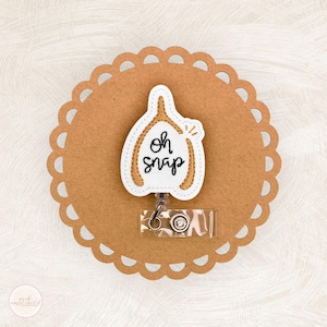 May include: A white and gold embroidered badge reel with the phrase "Oh Snap" in black script. The badge reel is shaped like a wishbone and is attached to a clear plastic retractable reel. The badge is on a brown scalloped circle.