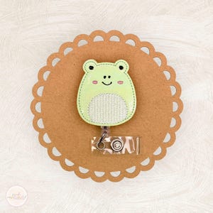 May include: A light green frog-shaped badge reel with a smiling face and a white mesh belly. The badge reel is attached to a clear plastic holder with a metal clip. The frog is on a brown, scalloped-edge background.