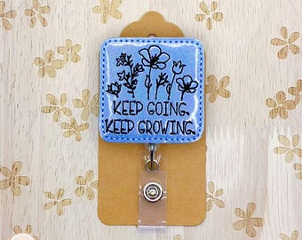 May include: A light blue badge reel with the words "KEEP GOING. KEEP GROWING." and a floral design. The badge reel is attached to a brown card with a clear plastic holder and a metal clip.