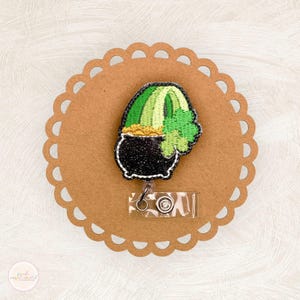 May include: A St. Patrick's Day themed retractable badge reel featuring a black pot of gold with a green shamrock and a green and yellow rainbow. The badge reel is attached to a scalloped brown card.