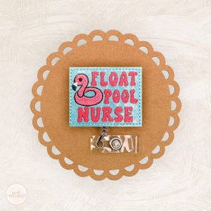 May include: A rectangular badge reel featuring light blue fabric with pink embroidered text that reads FLOAT POOL NURSE and a pink flamingo graphic. The badge reel has a clear plastic strap and a metal snap button attachment mechanism.