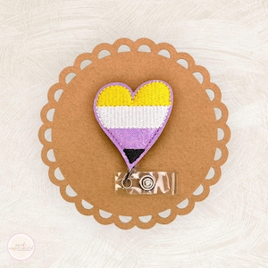 May include: A heart-shaped badge with the non-binary pride flag colors: yellow, white, purple, and black. The badge is on a brown, scalloped-edge paper circle. A clear plastic badge holder with a metal clip is attached to the bottom of the heart.