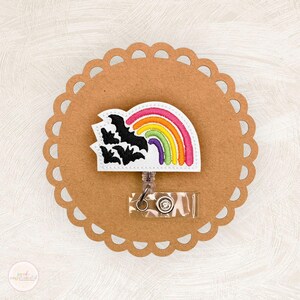 May include: A badge reel with a rainbow and bats design. The badge reel features a white background with a colorful rainbow and black bats. The reel is attached to a clear plastic holder with a metal clip.