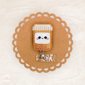 May include: A badge reel featuring a smiling pill bottle design with a glittery orange body and a white label. The badge reel is attached to a clear plastic holder with a metal clip, set on a brown, scalloped background.