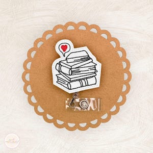 May include: A badge reel featuring a stack of books with a heart-shaped thought bubble. The design is white with black outlines, set on a brown, scalloped background. A clear plastic reel with a metal clip is attached.