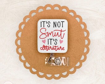 Può includere: Un porta badge bianco con la scritta "IT'S NOT Smut IT'S cliterature" in lettere nere e rosse, con due cuori rosa. Il badge è su uno sfondo marrone con bordi smerlati.
