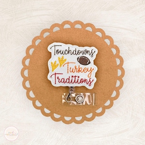 May include: A white embroidered badge with a scalloped edge featuring the text Touchdowns Turkey Traditions in brown, orange, and red thread. The design includes yellow bird footprints and a brown football. A clear plastic badge reel is attached.