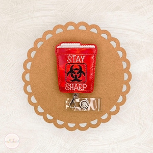 May include: A red badge reel with the words "STAY SHARP" and a biohazard symbol. The badge reel is in the shape of a sharps container. The reel is attached to a clear plastic holder with a metal clip.