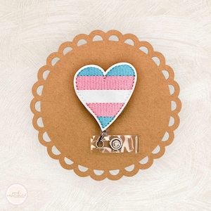 May include: A heart-shaped badge with the transgender pride flag colors: light blue, pink, and white. The badge is embroidered and attached to a clear plastic retractable reel with a metal clip. The badge is on a brown, scalloped-edge background.