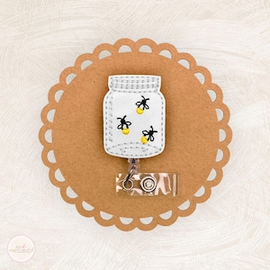 May include: White felt mason jar with a white felt lid and a white felt retractable badge reel. The jar is filled with fireflies with yellow bodies and black wings. The jar is on a brown background.