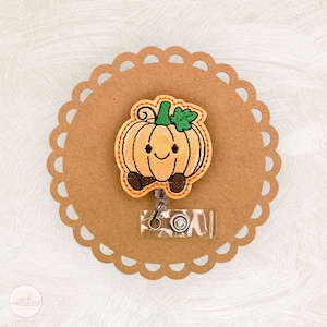 May include: A badge reel featuring a smiling pumpkin design with orange, green, and brown accents. The pumpkin is attached to a clear plastic reel with a metal clip, set on a scalloped brown paper background. Perfect for fall.